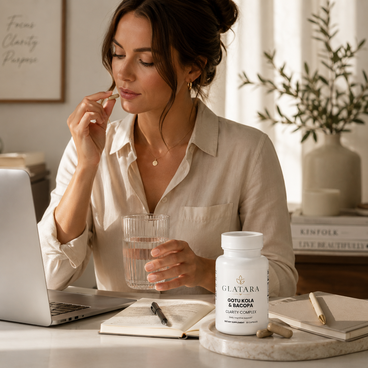 A woman in a beige button-up shirt sits at a desk, holding a glass of water and taking a supplement capsule from a white bottle labeled GLATARA SOOTY KOLA & BAMBOO. The scene is styled with a laptop, notebook, and a vase in the background.