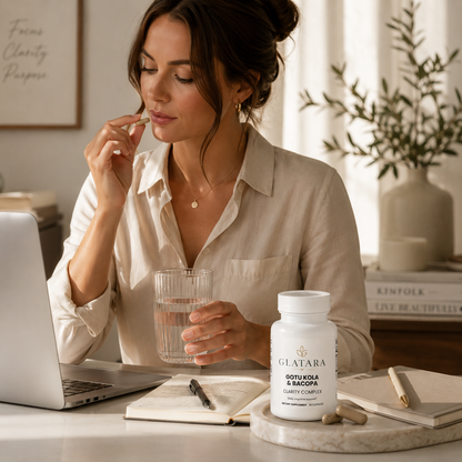 A woman in a beige button-up shirt sits at a desk, holding a glass of water and taking a supplement capsule from a white bottle labeled GLATARA SOOTY KOLA & BAMBOO. The scene is styled with a laptop, notebook, and a vase in the background.
